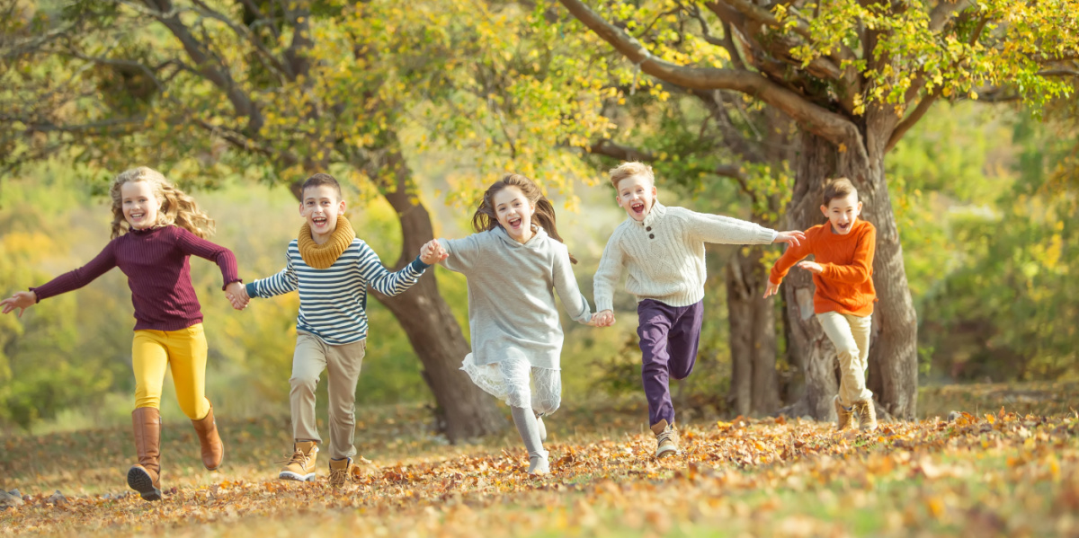 Fünf Kinder rennen über eine herbstliche Wiese, Im Hintergrund Bäume mit buntem Laub