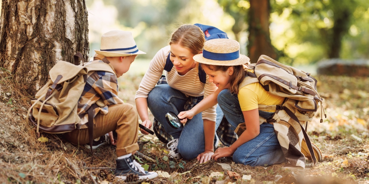 Zwei Kinder mit Hüten und eine Frau knien unter einem Baum und sammeln etwas