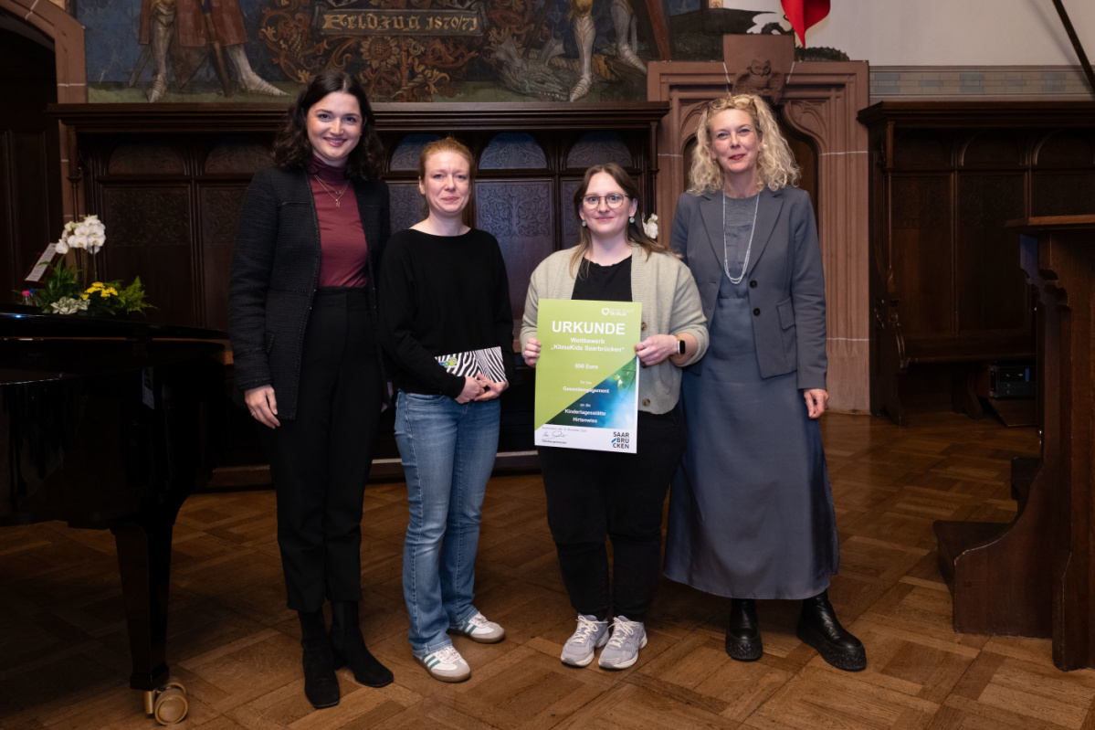 Preisübergabe an die Kita Hirtenwies. Zu sehen sind zwei Betreuerinnen der Grundschule die die Urkunde halten, sowie zwei Jurymitglieder.