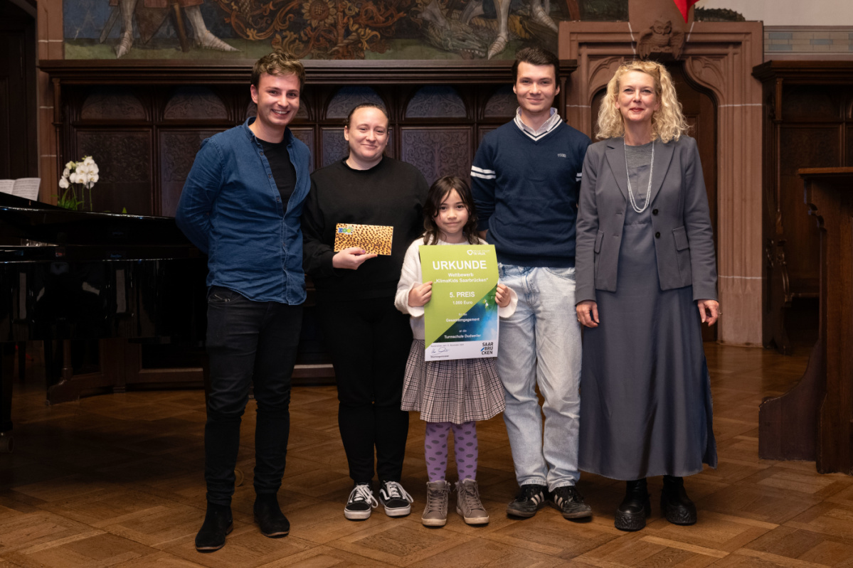 Preisübergabe des fünften Platzes an die Grundschule Dudweiler. Zu sehen ist eine Schülerin welche die Urkunde hält und zwei Betreuer der Grundschule, sowie zwei Jurymitglieder.