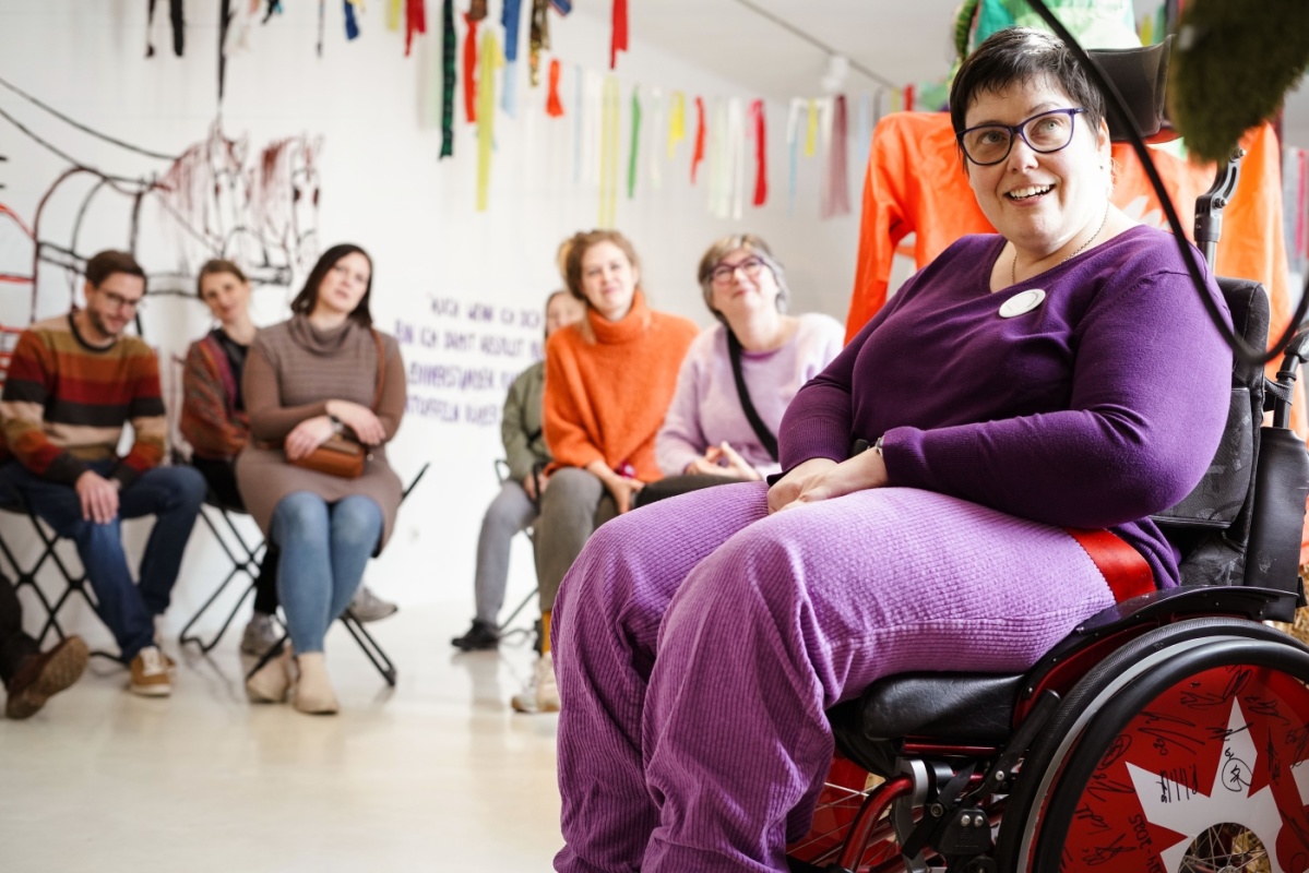 Frau in Rollstuhl sitzt vor einer Gruppe von Menschen, alle schauen begeistert in Richtung der Kamera aber an der Kamera vorbei