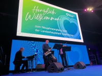 Eine Band mit vier Musikern auf einer Bühne, im Hintergrund eine Projektion mit der Aufschrift "Herzlich Willkommen zum Neujahrsempfang 2025"