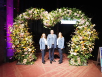 Drei Menschen in Malteser Hilfsdienst Uniform unter einem großen herzförmigen Blumen-Arrangement