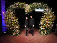 Zwei junge  Menschen in schwarz unter einem großen herzförmigen Blumen-Arrangement