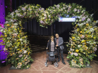 Ein Mann und eine Frau mit Rollator vor einem großen herzförmigen Blumenarrangement 