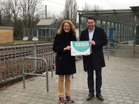 Zwei Personen stehen an einem Bahnhof und halten ein Schriftstück in den Händen