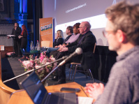 ein Mann steht am Laptop und auf der Bühne sitzen Menschen auf dem Podium